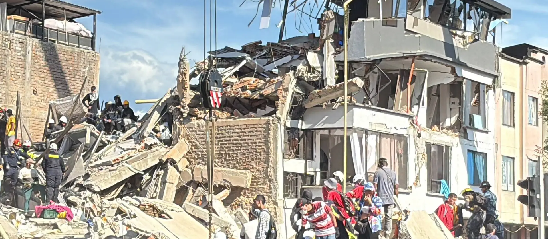 A multi-storey building in Pereira reduced to rubble, with rescue workers climbing the debris and residents gathered in the street below.