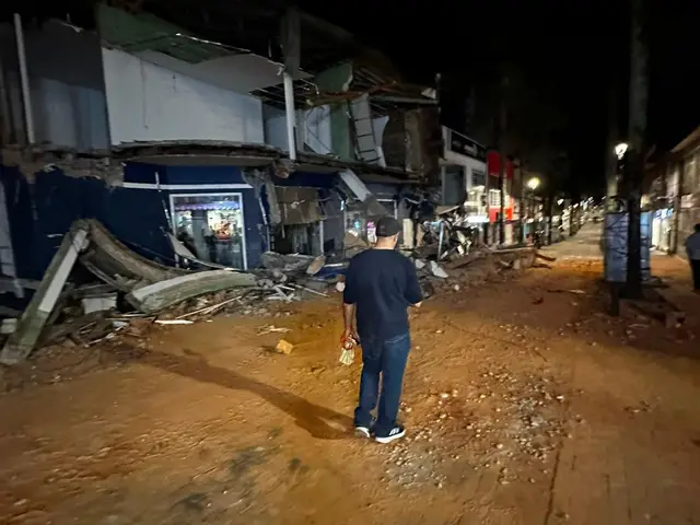 A person stands at night in front of shops with a collapsed facade and debris spilling across the road.